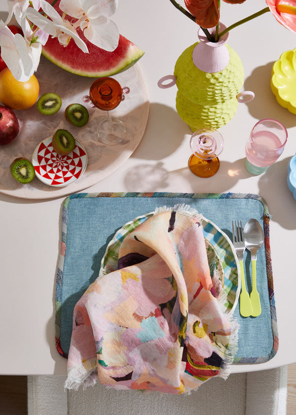 Colorful table setting with a floral napkin, cut fruits, and decorative items on a white table.