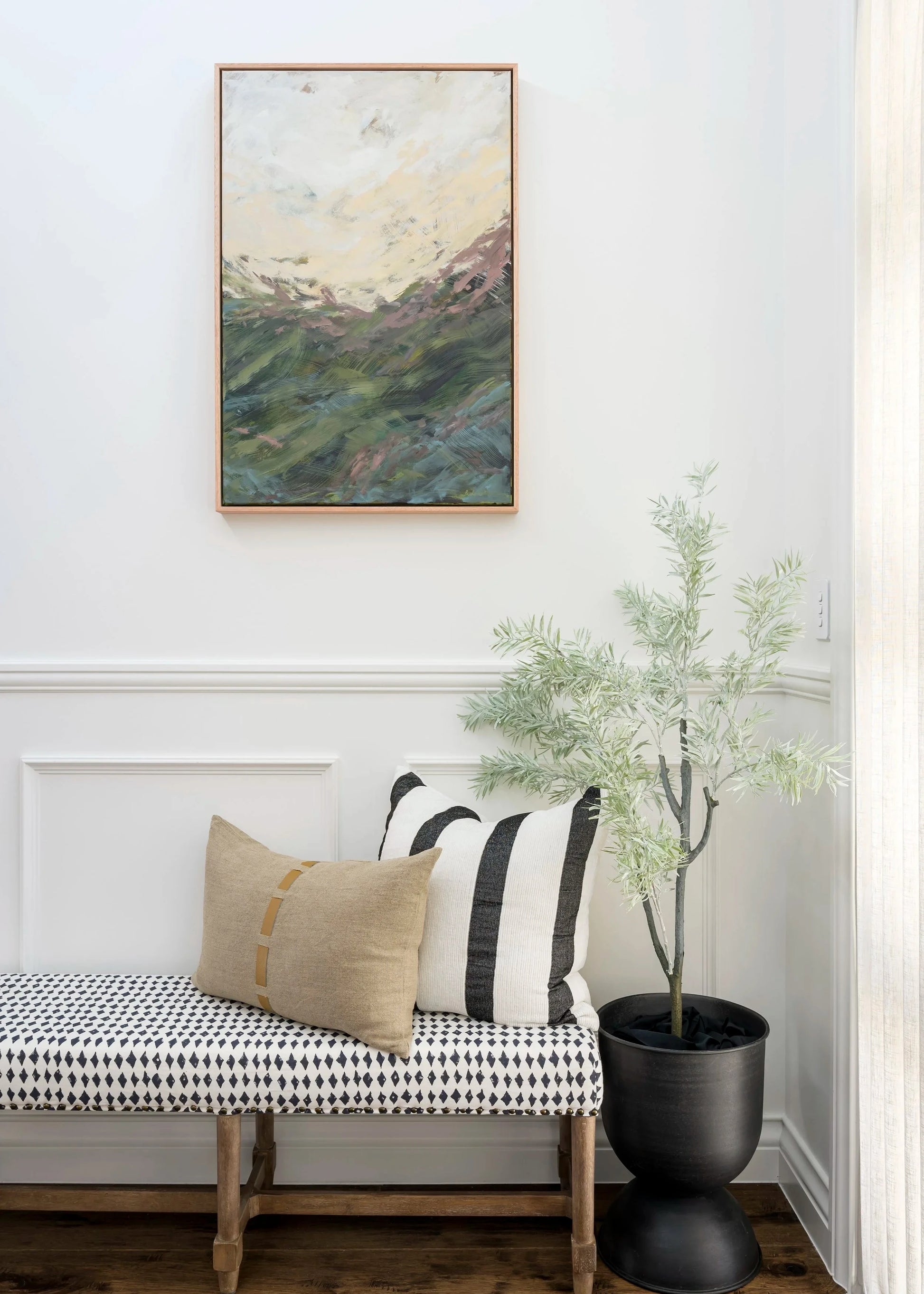 Narrow hallway with a bench, plant, and framed landscape painting on a white wall.
