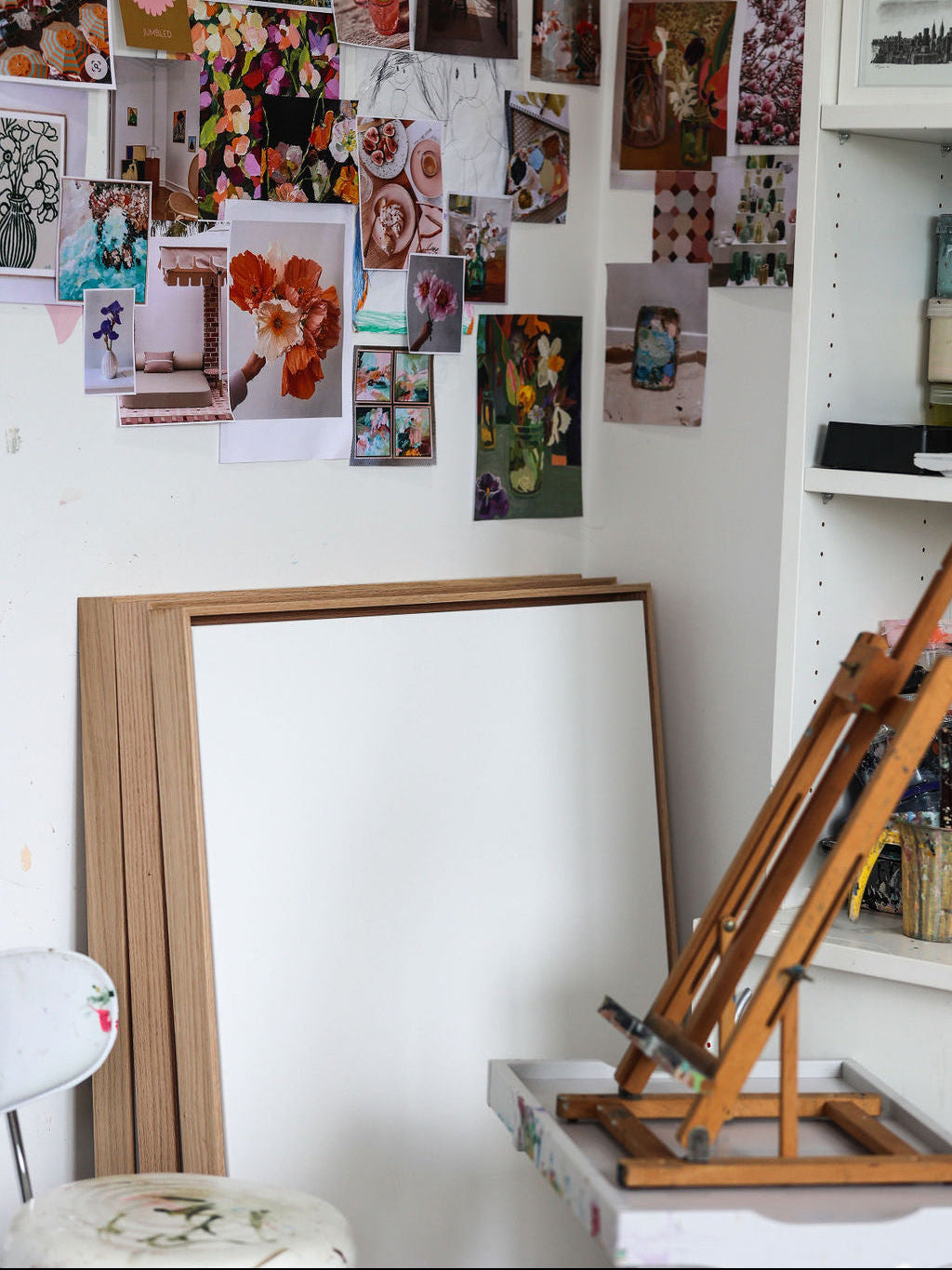 An artist's studio corner featuring a wooden easel, a large blank white canvas frame leaning against a wall, and a white pegboard covered with various art prints and sketches.